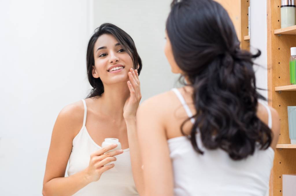 Queixas dermatológicas em cada fase da vida – pele jovem Woman caring of her beautiful skin on the face standing near mirror in the bathroom. Beautiful young woman applying moisturizer on her face. Smiling girl holding little jar of skin cream and applying lotion on face.