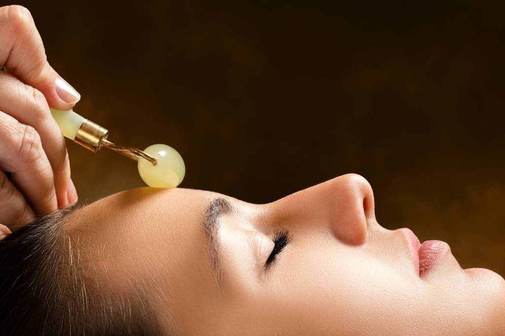 Técnicas e instrumentos de massagem, como o role de jade Therapist applying jade roller on female face.