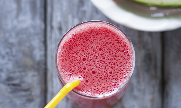 receita-suco-calmante Um copo de vidro com suco de cor vermelha e canudo amarelo aparecem sobre um fundo de madeira acinzentado