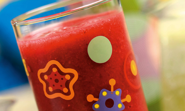 receita-suco-de-amora-com-morango-e-agua-de-coco Um copo de vidro com desenhos coloridos cheio de suco de cor vermelha aparece sobre fundo desfocado azul e verde