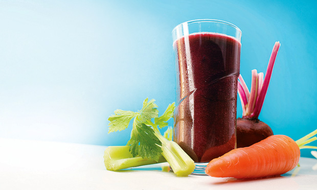 receita-suco-de-beterraba-cenoura-salsao-e-erva-doce Um copo de vidro cheio com suco de cor roxa é cercado por dois pedaços de salsão, uma cenoura e uma beterraba