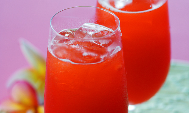receita-suco-flores-frutas-2 Dois copos de vidro com gelo e suco de cor vermelha aparecem no primeiro plano sobre uma superfície verde. O fundo é roxo com pedaços de flores aparecendo