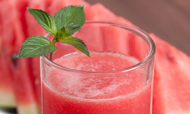 receita-suco-melancia-boa-forma copo de suco de cor vermelha com folhas de hortelã para decorar. Ao fundo, aparecem fatias de melancia desfocadas