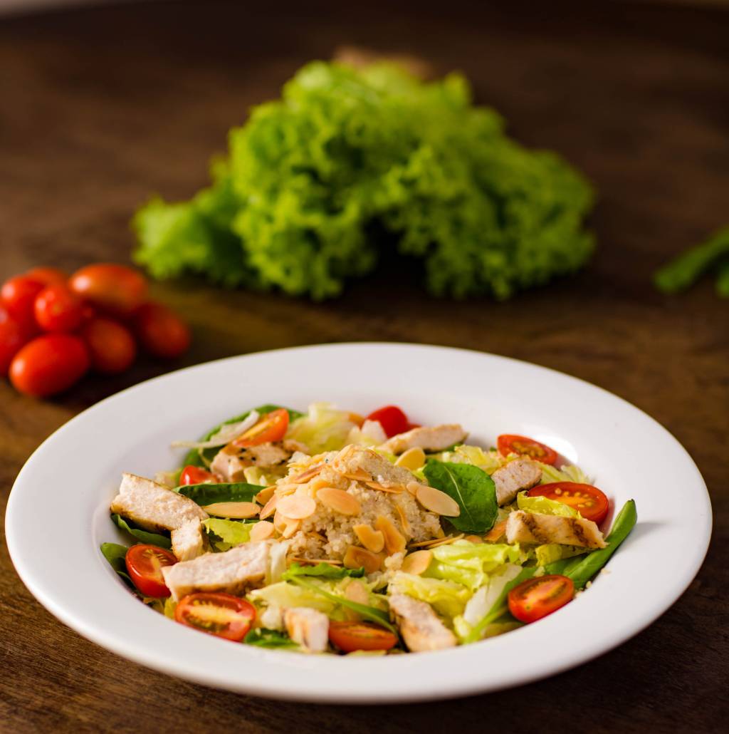salada-proteica-restaurante-madero Em um prato branco fundo, há uma salada com folhas verdes, tomates, pedaços de frango e quinoa. No fundo, desfocado, aparece um pé de alface e tomates-cereja