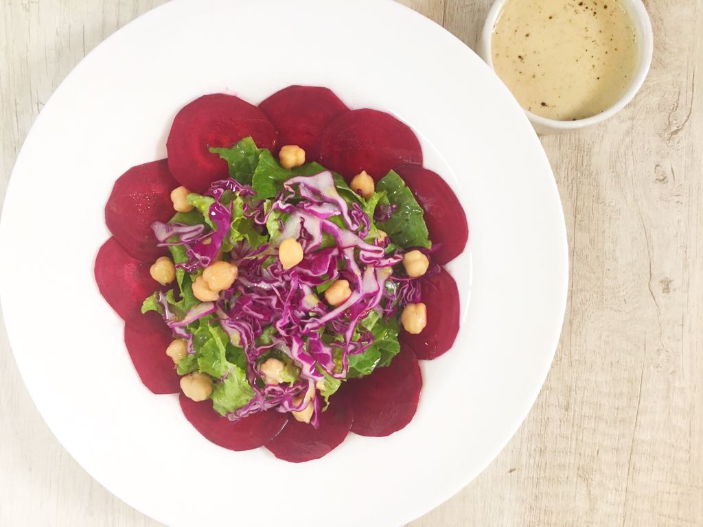 salada-carpaccio-beterraba-grao-de-bico-tahine Em um prato branco aparecem diversas fatias de beterraba cortadas bem finas, como um carpaccio. No meio, uma salada de grãos, folhas verdes e repolho
