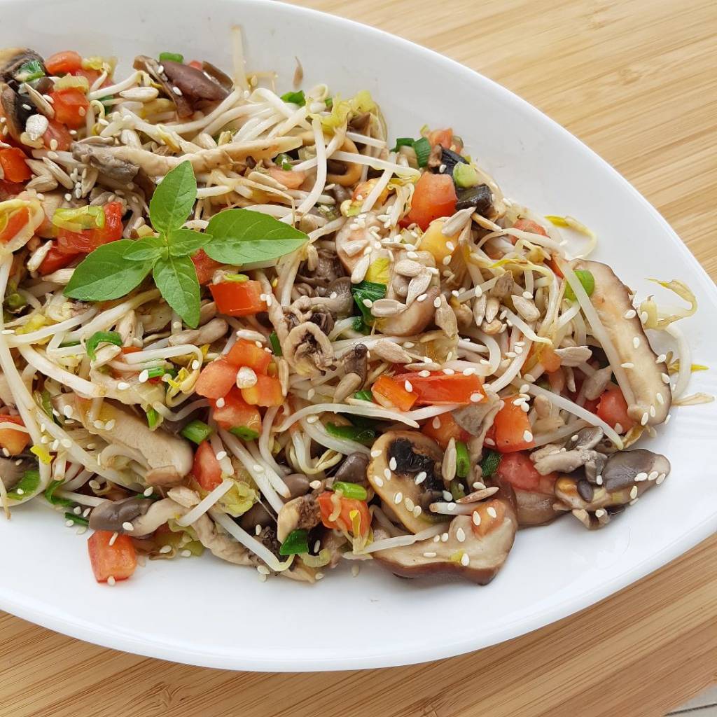 salada-quente-broto-feijao-cogumelos Em uma travessa branca de louça, misturam-se broto de feijão, cogumelos e pedaços de tomate. Uma folha de manjericão decora o prato, que está sobre uma superfície de madeira.
