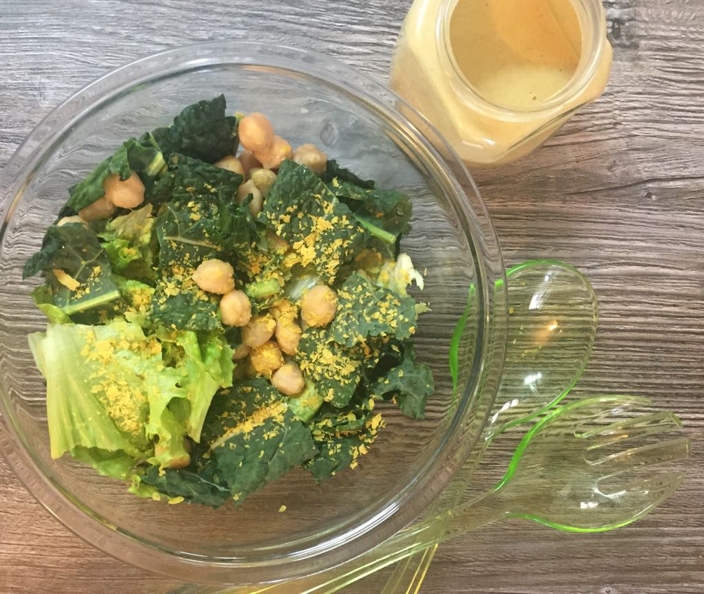 salada-couve-grao-de-bico-tahine Em uma tigela de vidro, há uma salada de folhas verdes com grão de bico. Ao lado, há um pote com molho de tahine. Sobre a superfície de madeira, há duas colheres de plástico esverdeadas