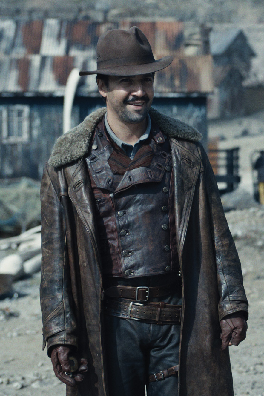 Homem com roupas de couro e chap&eacute;u caminha em cen&aacute;rio de fazenda antiga