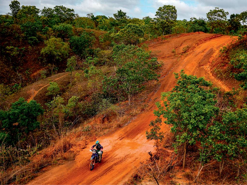 Moara-sacilotti-rally-dos-sertoes Uma faixa aberta em meio à vegetação vira pista no cenário de terra, com chão vermelho. Uma moto, pilotada por uma mulher, aparece no centro da pista