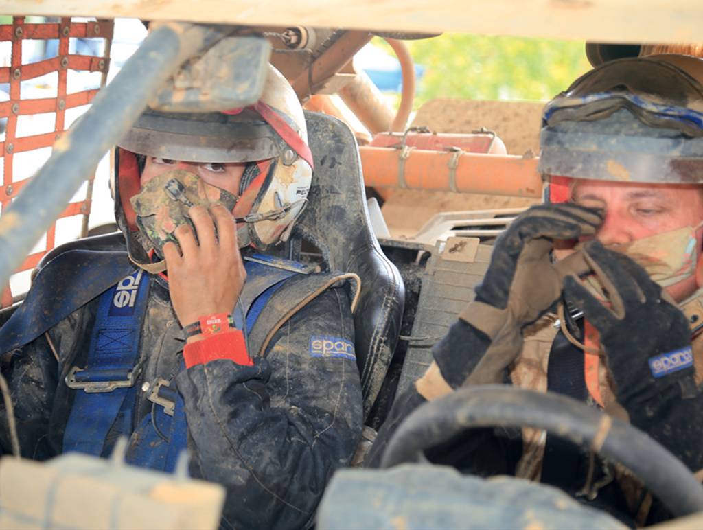 corina-neumann-nicole-neumann-rally-dos-sertoes Duas mulheres estão sentadas nos bancos da frente do veículo. Elas estão com o uniforme, máscara e capacete, então só é possível enxergar os olhos delas. Elas e a superfície do carro estão sujas de terra e poeira levantadas na corrida.