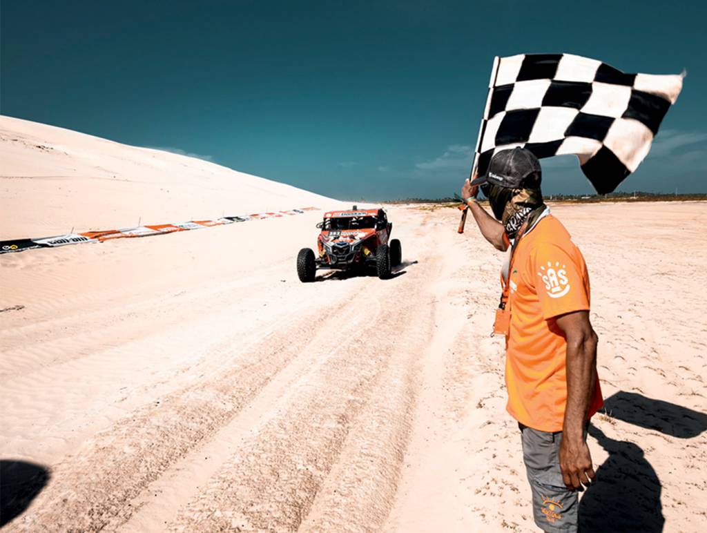 bandeirada-rally-dos-sertoes Em um terreno arenoso bem claro, um carro corre e se aproxima da bandeirada, dada por um homem de camiseta laranja