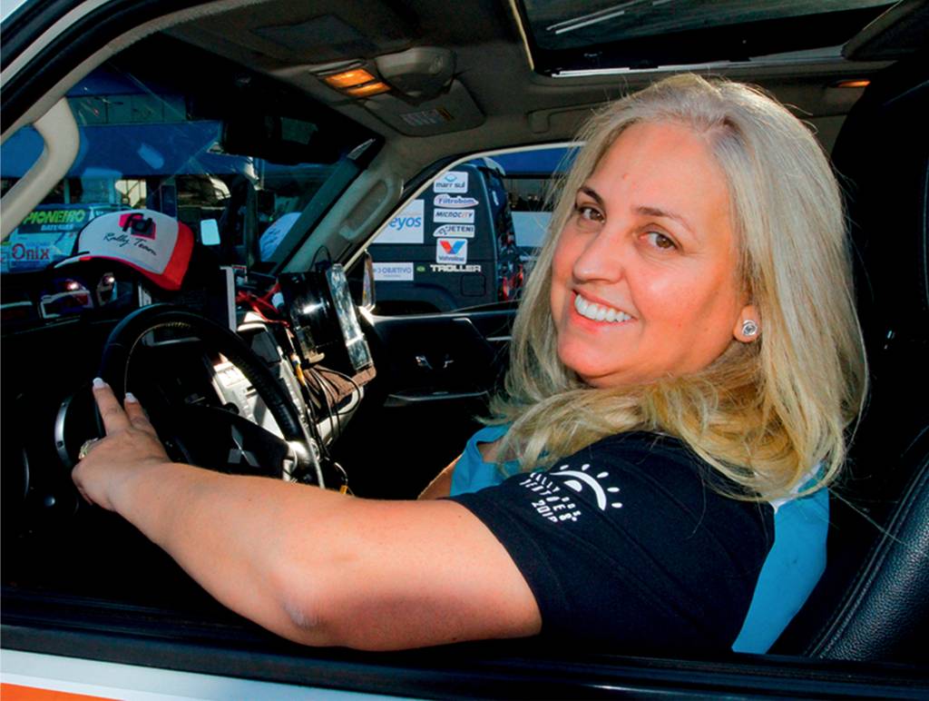 Sandra-Dias-rally-dos-sertões Mulher branca com cabelos loiros sorri para a foto. Ela está sentada dentro de um carro e com o braço apoiado na janela aberta.