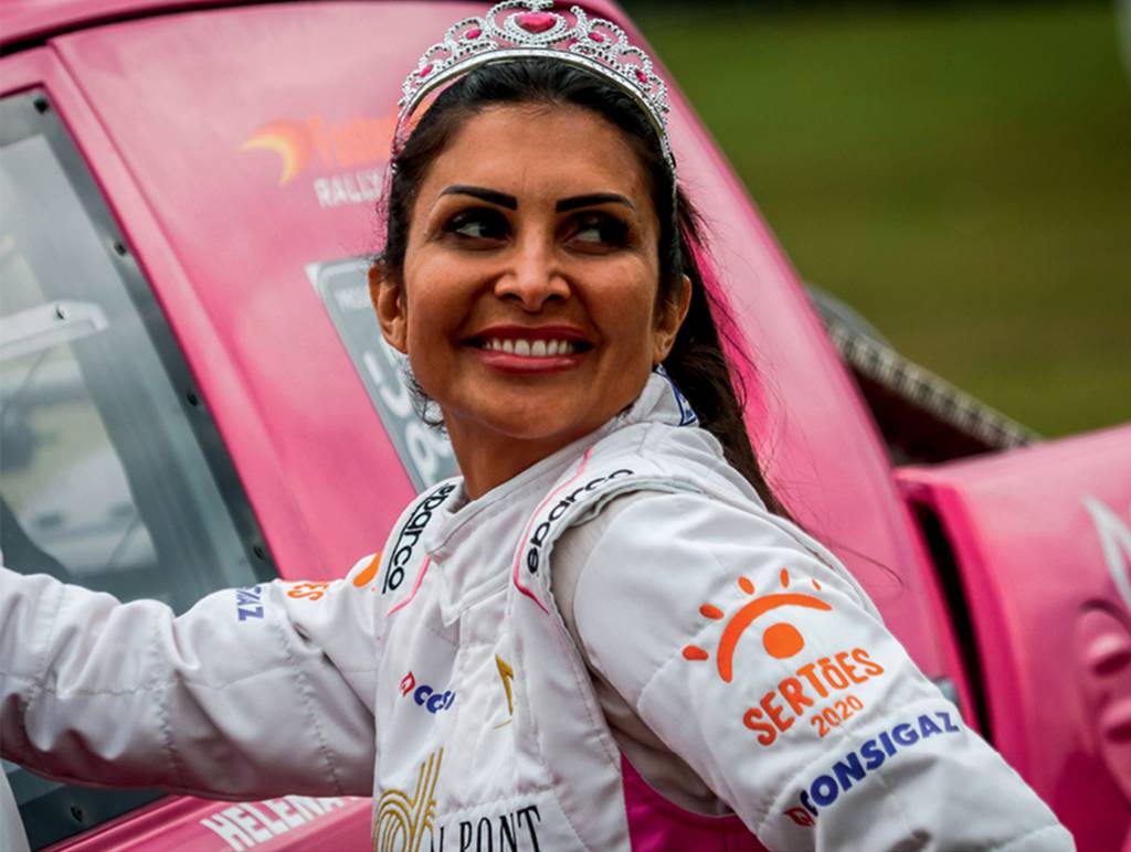 Helena-soares-rally-dos-sertoes Uma mulher de cabelos escuros presos sorri para a foto. Ela usa um uniforme de corrida e uma coroa. Está apoiada em sua caminhonete pink