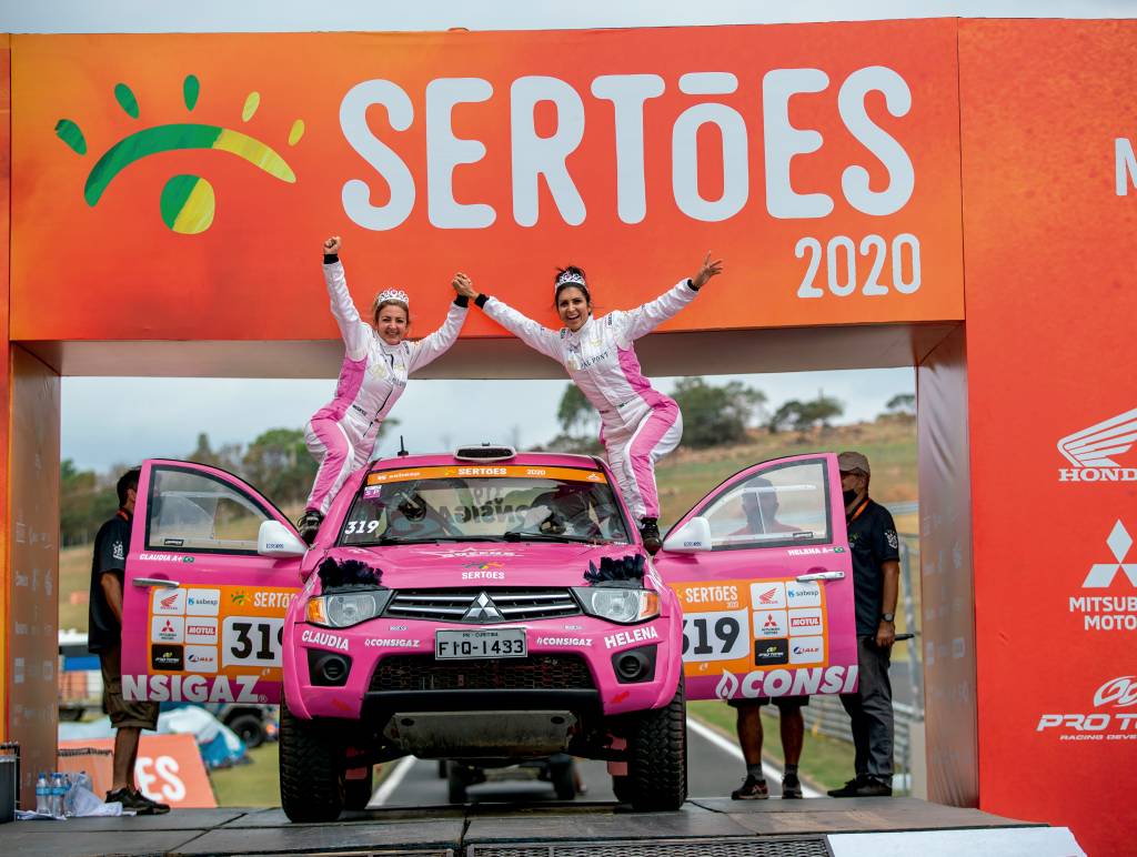 claudia-helena-rally-dos-sertões duas mulheres estão de pé apoiadas nas portas abertas de um carro cor de rosa. O farol do carro tem um item que imita cílios
