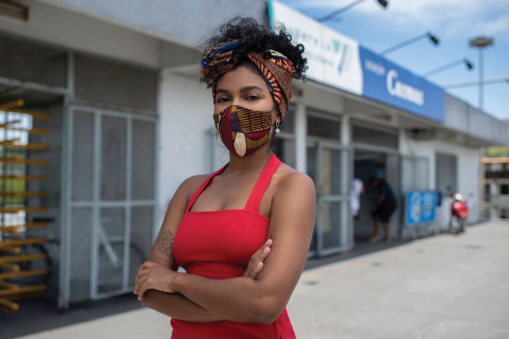 joyce-trindade Joyce usa macacão vermelho, lenço no cabelo e máscara no rosto. Ela está com os braços cruzados em frente a uma instituição pública