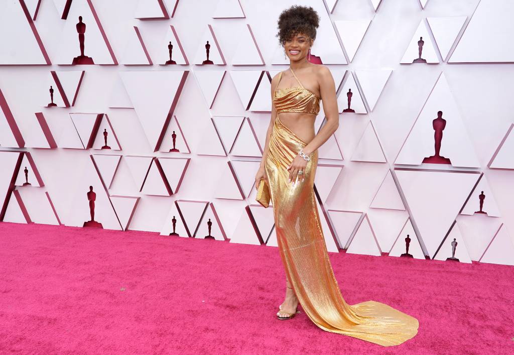93rd Annual Academy Awards – Arrivals Andra é uma mulher negra, com cabelos castanhos cacheados presos. Ela usa um vestido dourado com recorte abaixo do seio e por toda a lateral do torso.