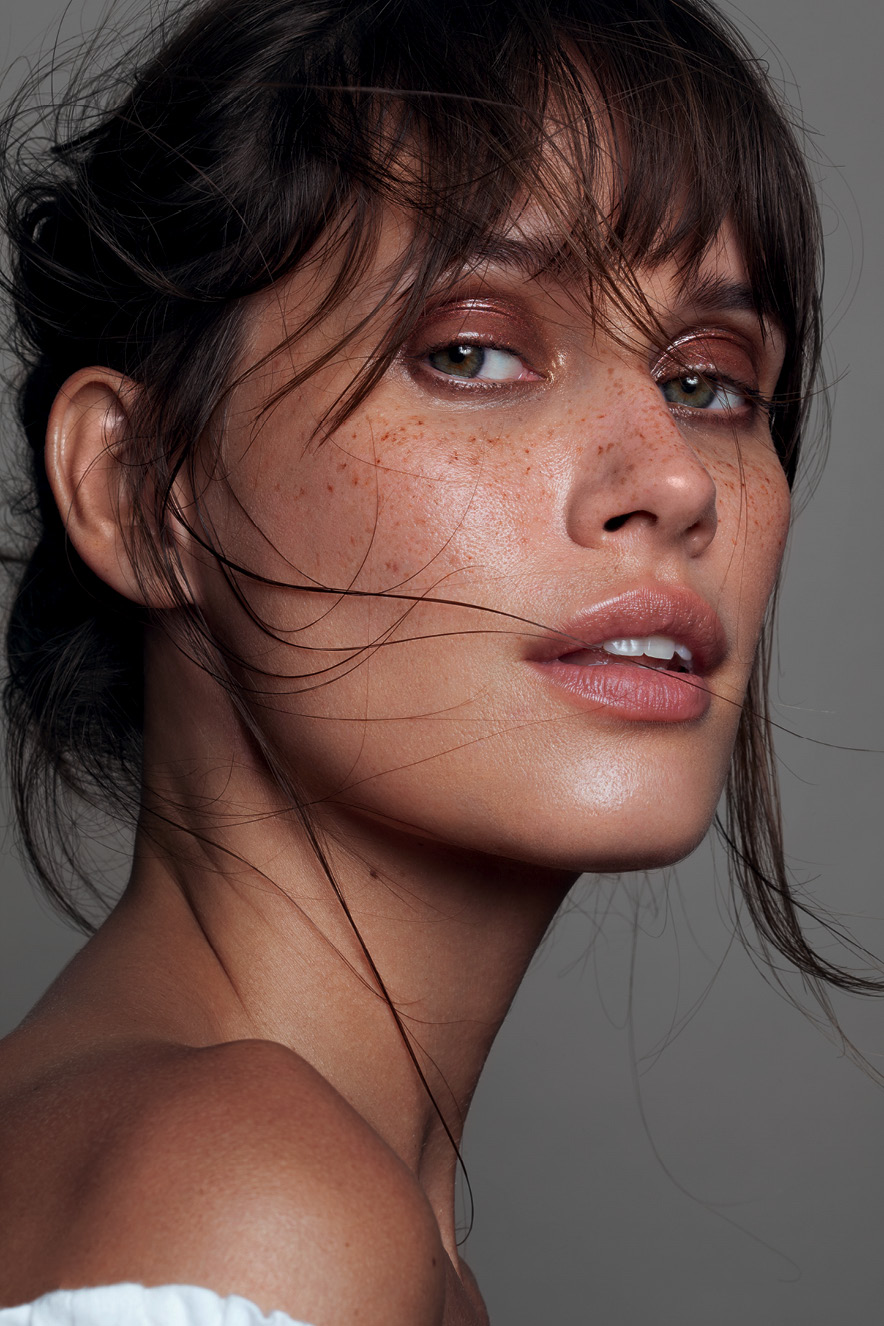 Em retrato, mulher maquiada com pele natural e olhos marcados por sombra marrom olha para a c&acirc;mera