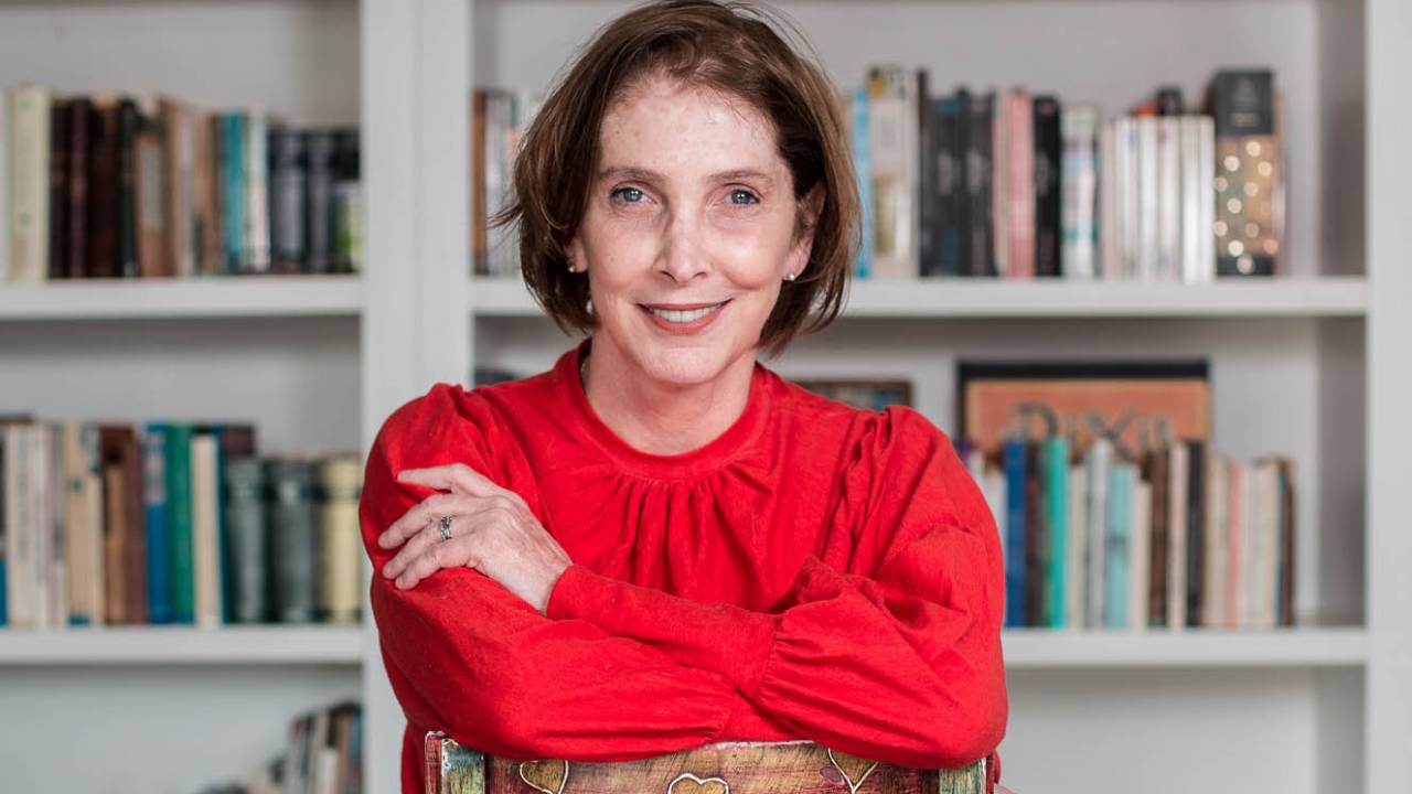 Adriana Falcão está sentada à frente de uma prateleira com livros
