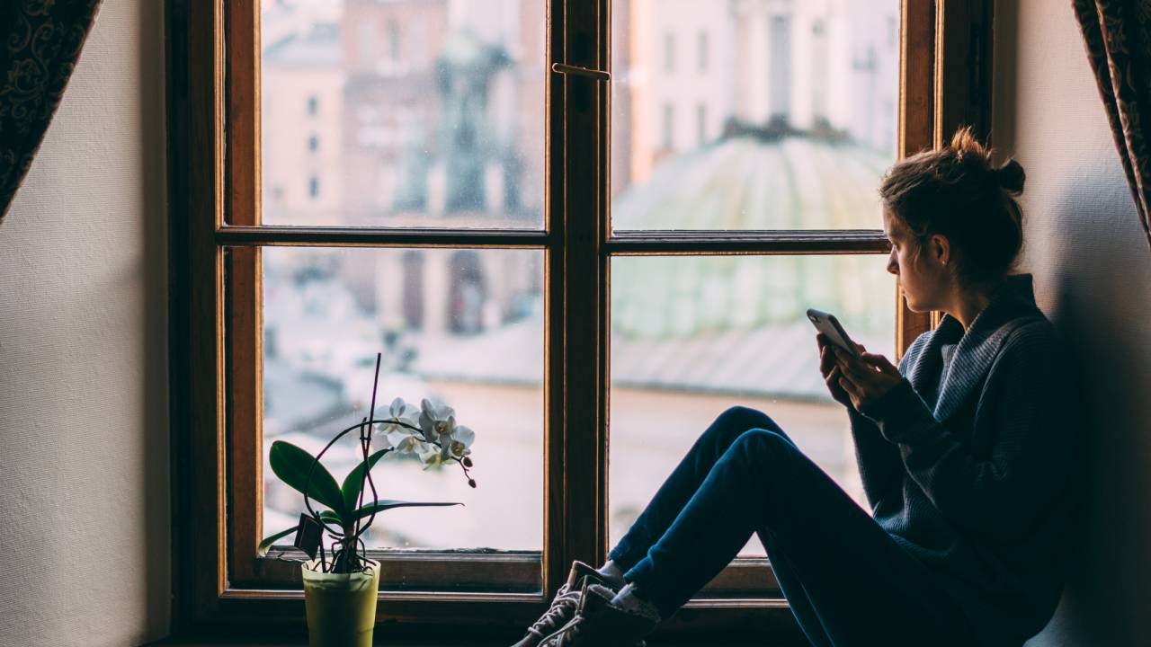 mulher olha pela janela e segura um celular