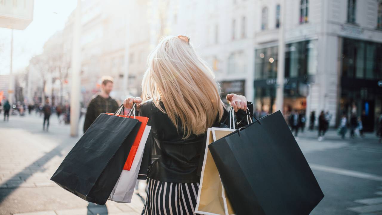 Mulher loira caminhando na rua com sacolas de compras nas mãos.