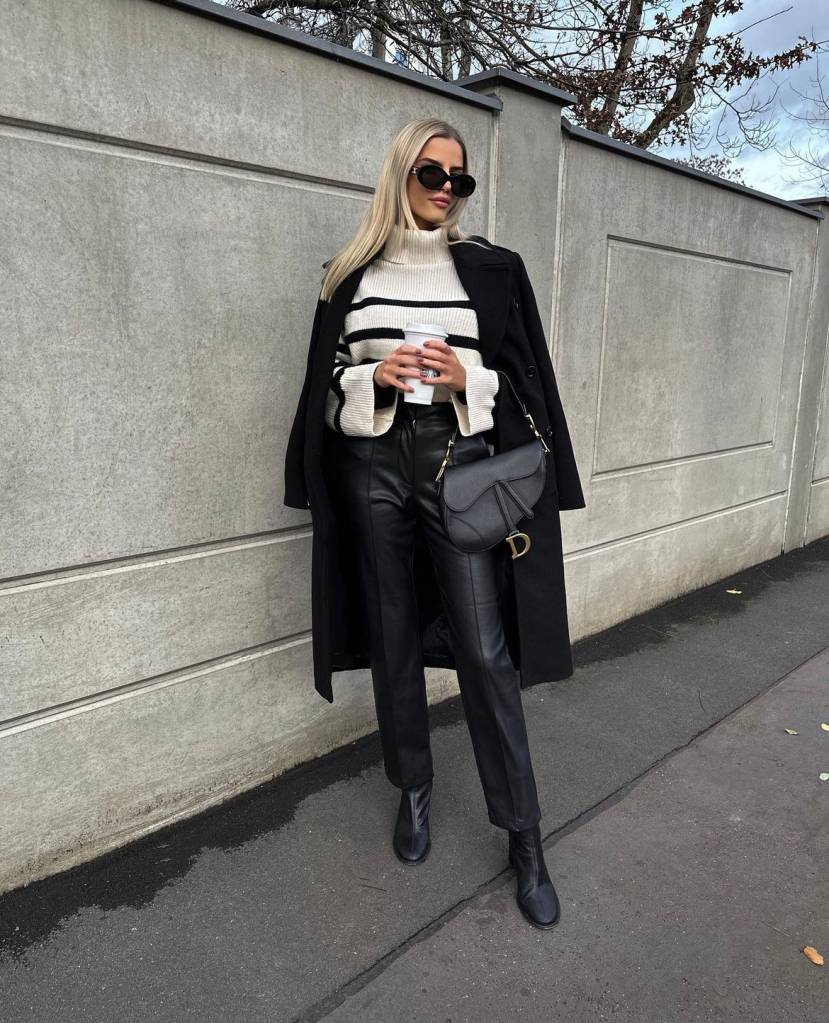 moda-outono-tendências-dicas-cores-2 A imagem mostra um mulher loira, segurando uma bolsa preta de couro e um copo de café/ Ela veste um suéter creme e preto listrado, com uma calça preta de couro com botas no mesmo material.