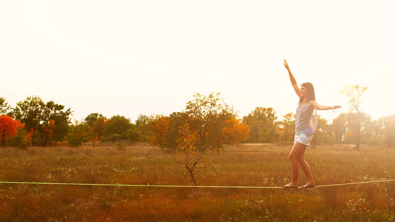 Mulher andando no slackline no prado ao pôr do sol. Indicação de misto de emoções como frio na barriga, medo e entusiasmo.