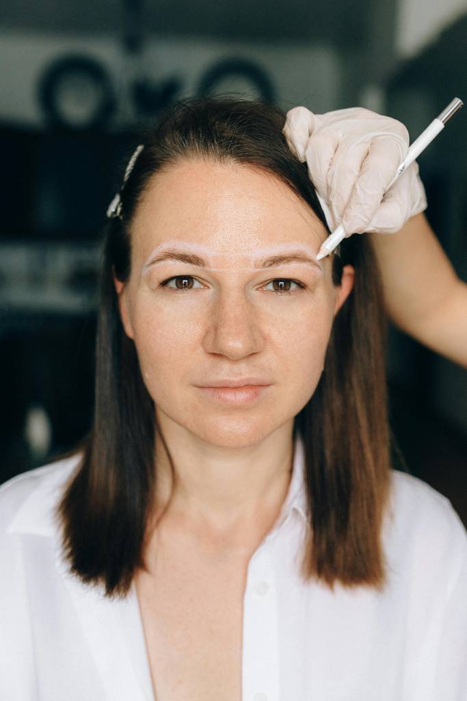 Imagem de mulher posando em frente a câmera enquanto é analisada por um profissional estético. Ela está sendo submetida a métricas de sua sobrancelha.