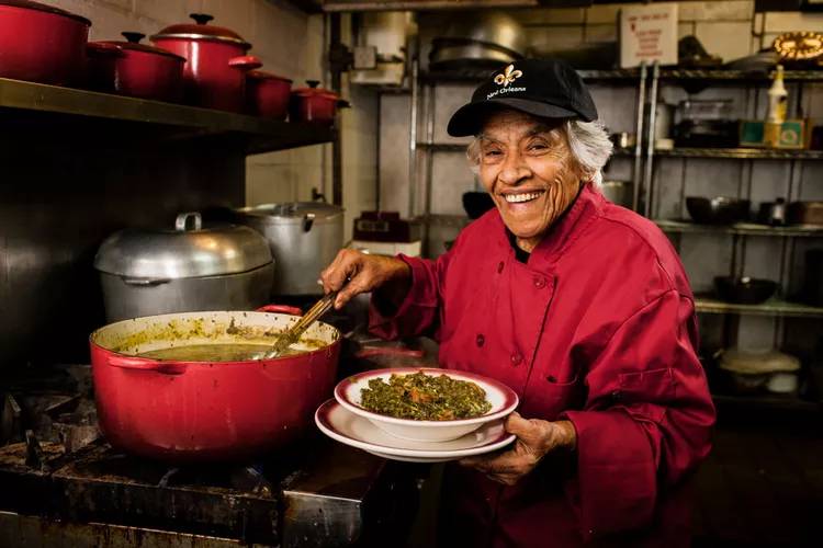 Imagem de uma pessoa idosa pegando a comida de uma panela e colocando em um prato em um dos restaurantes citados na matéria
