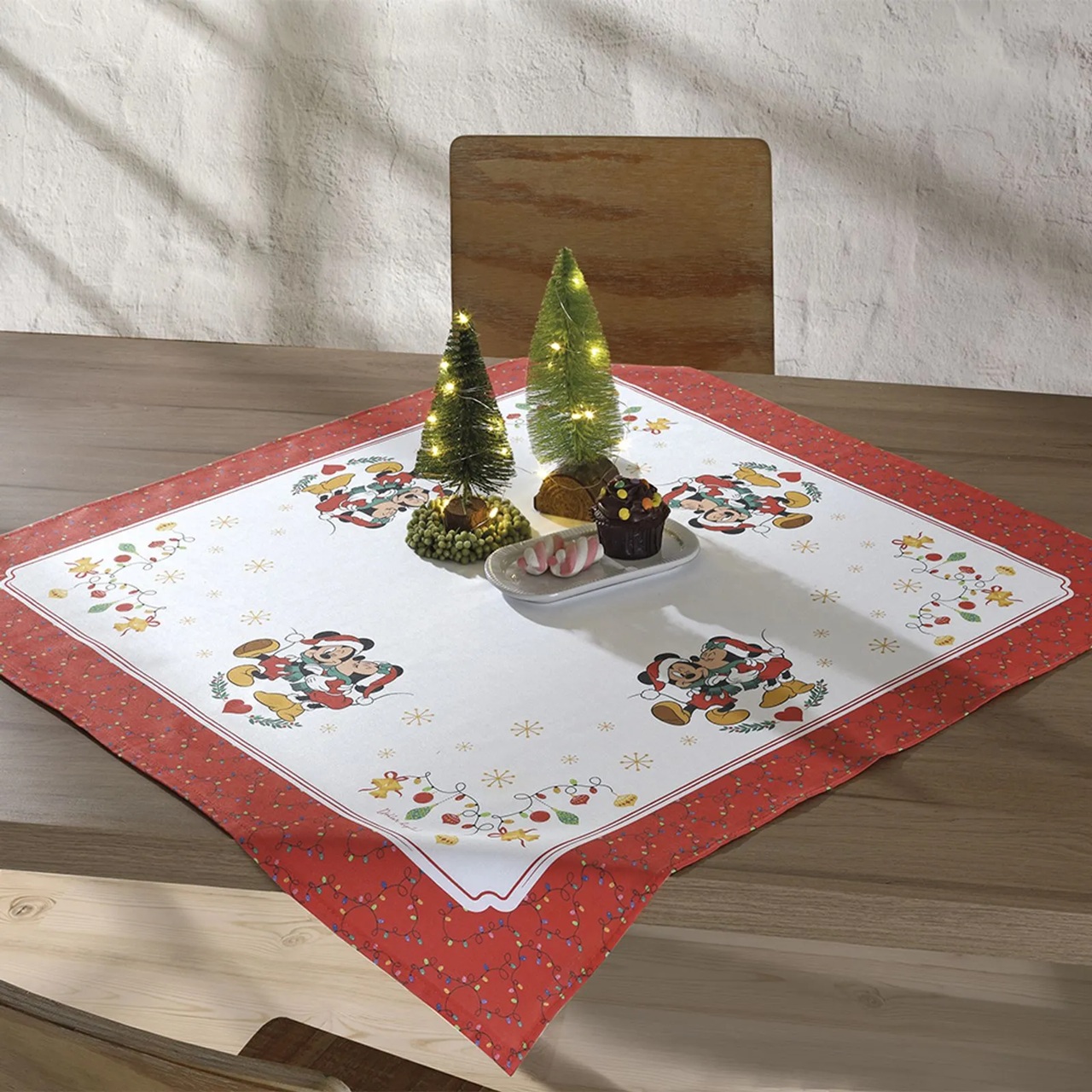 Toalha de Natal licenciada do Mickey e Minnie sobre a mesa, decorada com mini&aacute;rvores iluminadas e doces, item vendido nas Lojas Mel em S&atilde;o Paulo. A rede &eacute; conhecida por op&ccedil;&otilde;es econ&ocirc;micas de decora&ccedil;&atilde;o natalina, incluindo velas eletr&ocirc;nicas e pe&ccedil;as de mesa posta