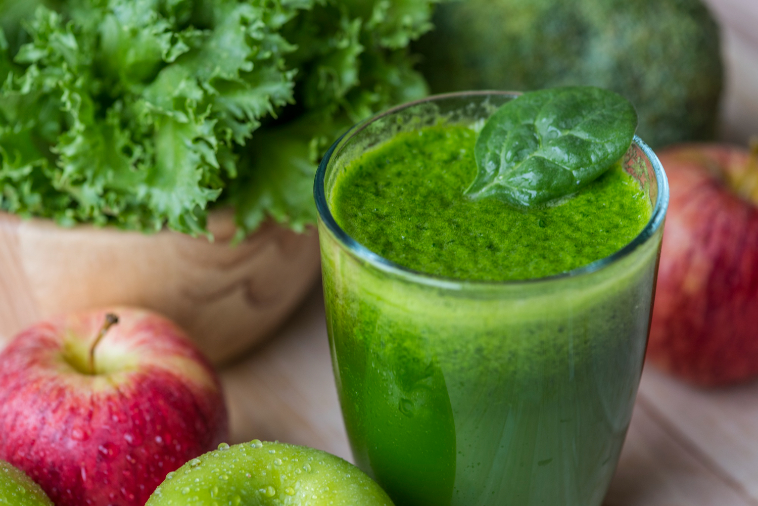 Shot de couve, ma&ccedil;&atilde; e lim&atilde;o em copo de vidro, com uma folha verde no topo. Ma&ccedil;&atilde;s vermelhas e verdes e um punhado de couve fresca est&atilde;o dispostos ao redor, destacando os ingredientes do suco verde para fortalecer a imunidade, rico em vitaminas A, C e zinco.