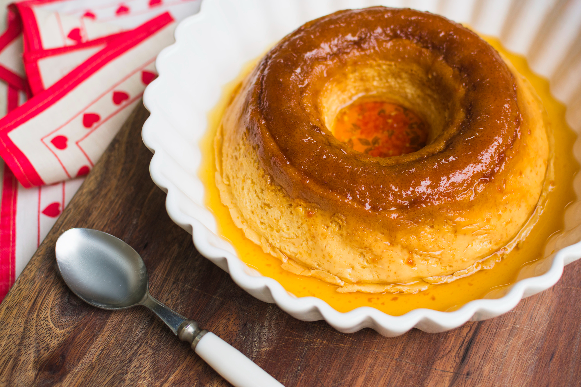 Pudim com calda de caramelo em formato de anel, servido em um prato branco com borda ondulada. Ao lado, h&aacute; uma colher e um pano decorado com cora&ccedil;&otilde;es vermelhos sobre uma mesa de madeira.