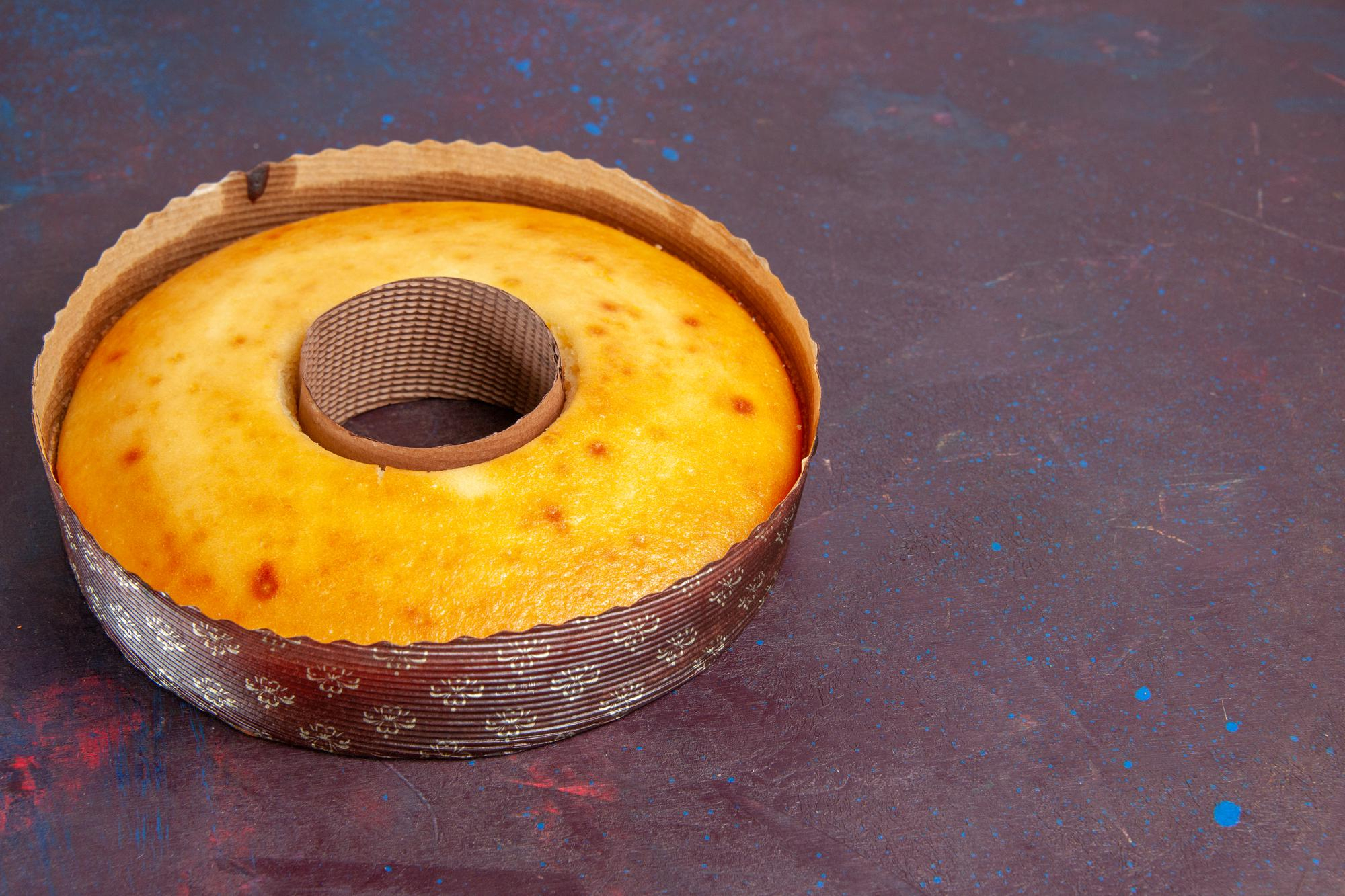 Bolo de milho redondo e dourado, assado em uma forma de papel com furo central, sobre uma superf&iacute;cie escura com respingos azulados, destacando o tom amarelo vivo da massa.