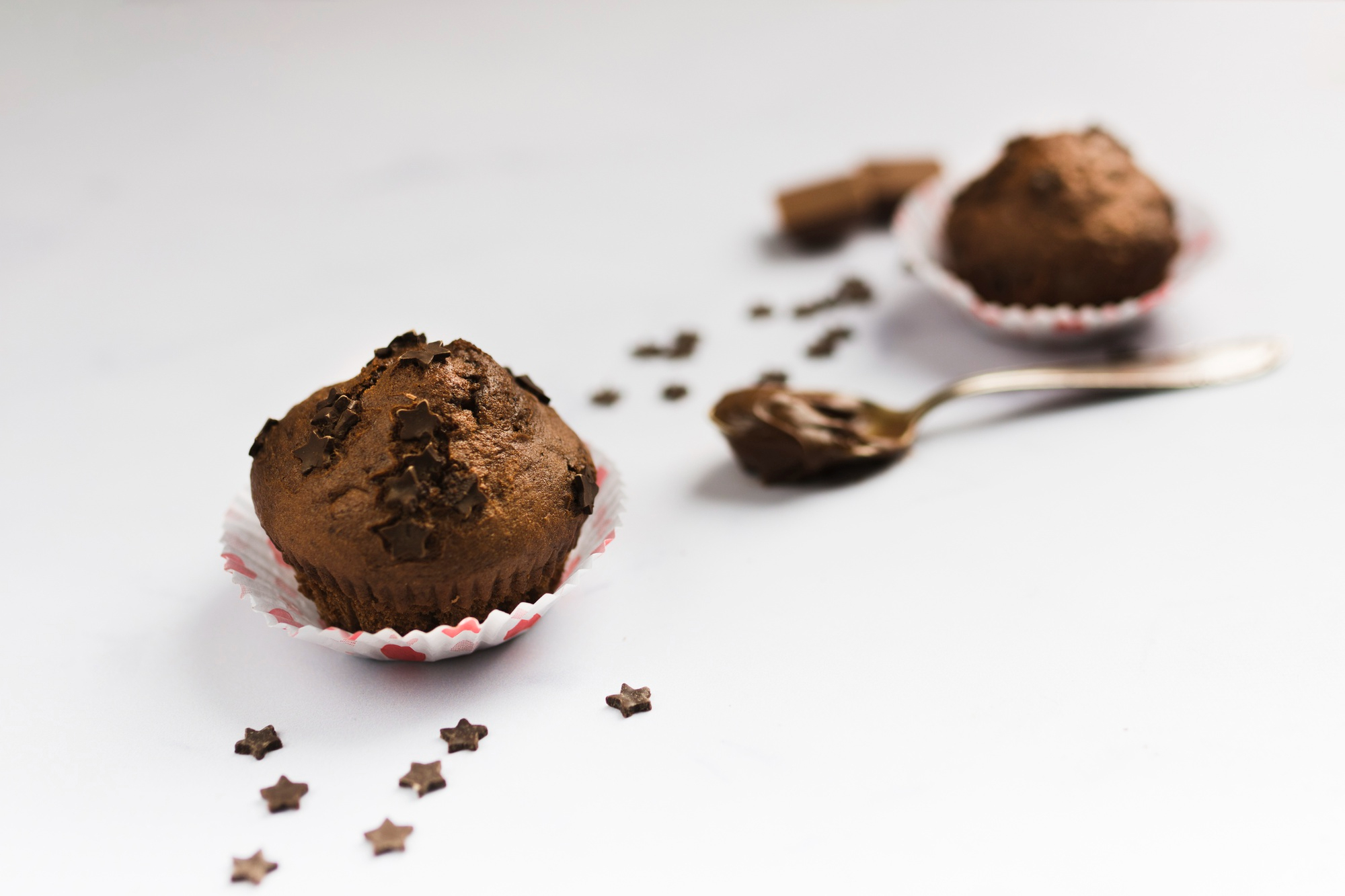 Cupcake de chocolate com confeitos em formato de estrelas no topo, em forminha decorada. Ao fundo, aparecem outro cupcake desfocado, uma colher com chocolate e pequenos confeitos espalhados sobre fundo branco.