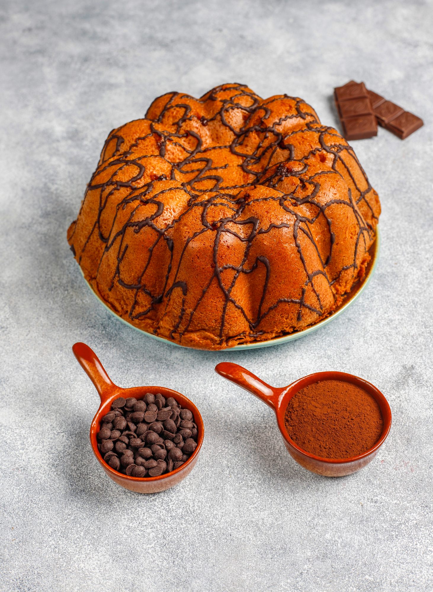 Bolo grande de cenoura em formato de pudim, com cobertura em fios de chocolate derretido por cima. Ao redor, aparecem duas colheres com gotas de chocolate e cacau em p&oacute;, al&eacute;m de peda&ccedil;os de chocolate ao fundo, sobre uma superf&iacute;cie cinza clara.