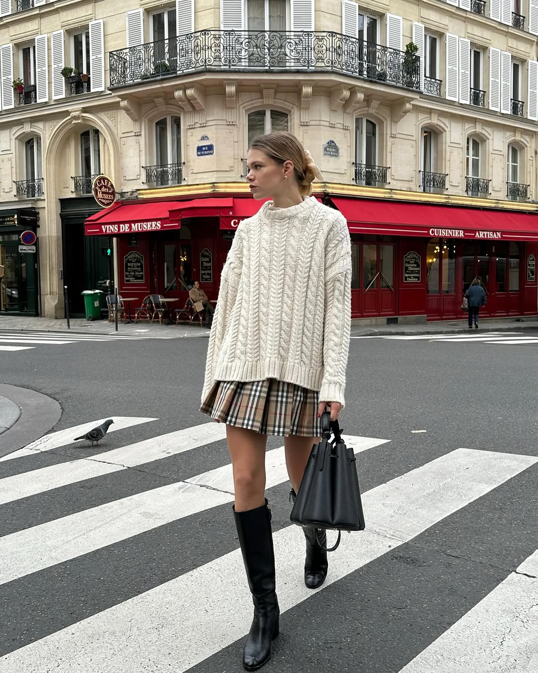 Foto de uma mulher loira usando um look composto por blusa de tric&ocirc; branca, saia plissada com listras, bota de couro e bolsa